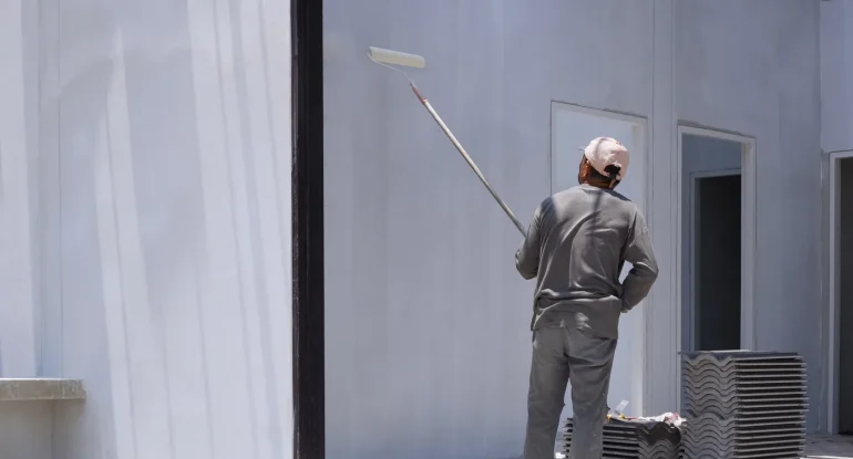 Trabajador pintando una pared blanca con rodillo, usando ropa de protección y un sombrero, en un entorno de construcción.